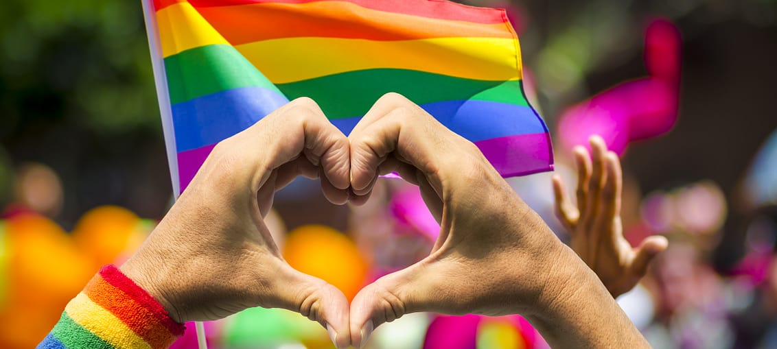 Pride flag (rainbow colours) with two hands in front of it. The hands are shaped into a heart.